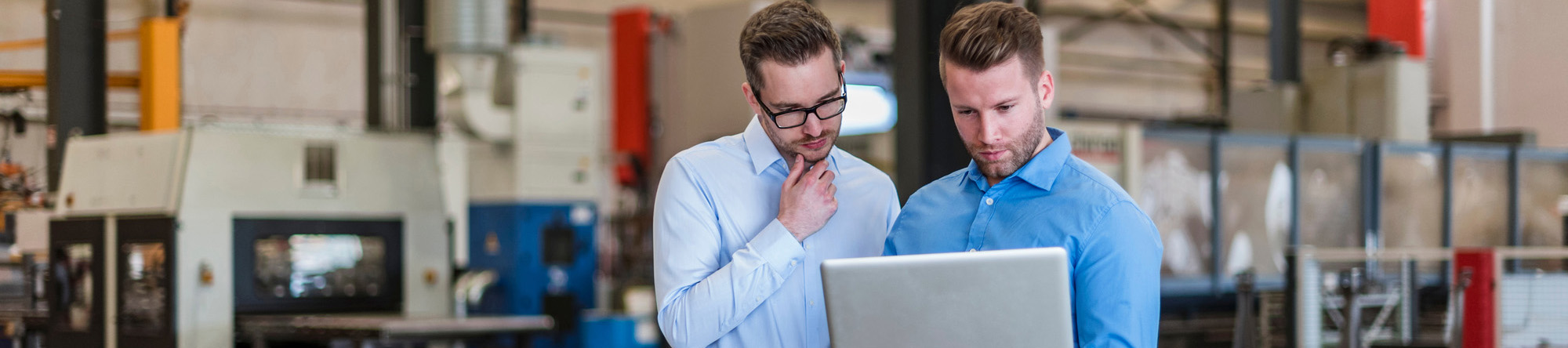 two people in an industrial setting looking at a laptop with machinery in the background