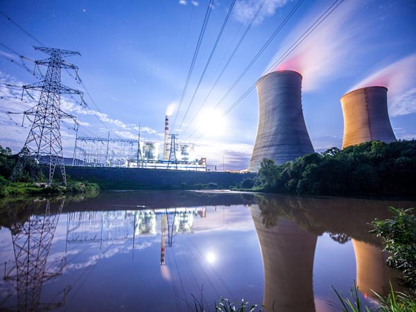 Power plant with cooling towers and electrical infrastructure by a reflective lake at dusk.