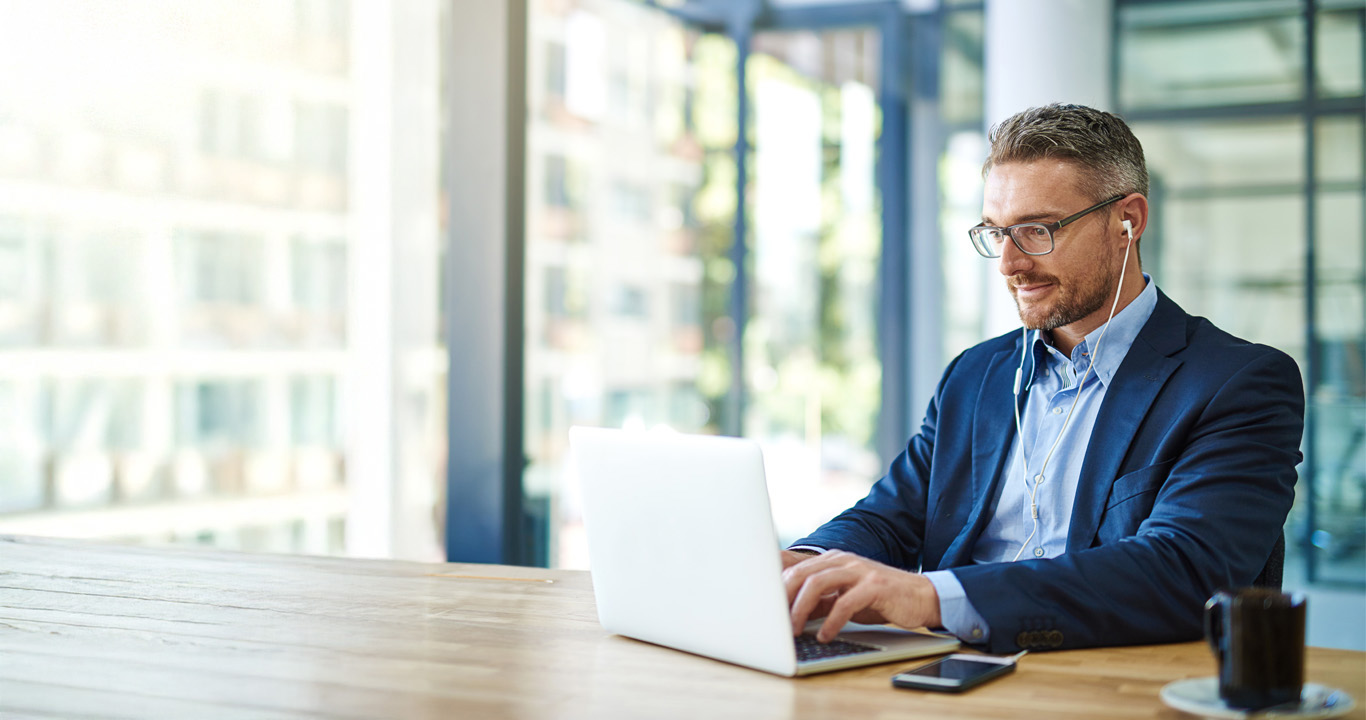 businessman working on a laptop in a modern office with large windows, wearing earphones and sitting at a wooden table with a smartphone and coffee cup nearby