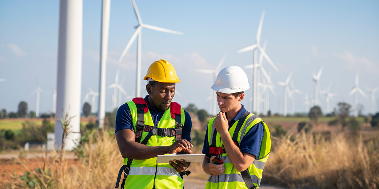 Two workers consult a tablet while standing in front of turbines at a wind farm. 
