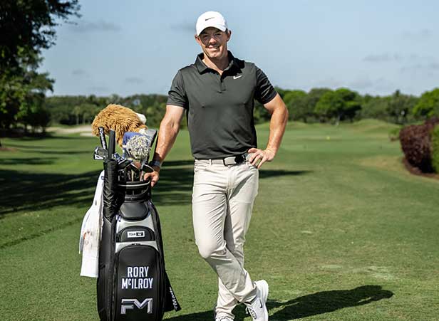 Rory McIlroy standing with golf bag, professional golf gear, outdoor sports activity, FM branded golf equipment, promoting wellness through golf, golf course landscape, enhancing sports lifestyle, golf attire and accessories, personalizing sports equipment, golfer ready for game